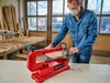 A man in a workshop is using a red scroll saw to cut a piece of wood. The workshop is filled with wood and tools. Produktbild-Vorschau