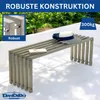 A metal garden bench with a hat and a book on it, set against a backdrop of greenery and a white wall. Produktbild-Vorschau