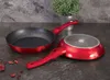 Two red frying pans are displayed on a dark, textured surface, with a potted plant in the background. Náhled obrázku produktu