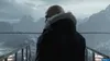 A bald man in a black winter coat with a fur-lined hood stands on a bridge overlooking a snowy mountain range. Produktbild-Vorschau