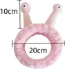 A pink, circular cosmetic headband with two antennae-like protrusions, resembling a snail. Dimensions are indicated by measurement lines and text. Produktbild-Vorschau