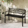 A black metal garden bench sits on a tiled patio, with plants and shadows from foliage visible in the background. Podgląd zdjęcia produktu