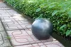 A dark gray garden sphere sits on a patterned tiled pathway next to a dense green hedge. Produktbild-Vorschau