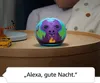 An Amazon Echo Dot Kids with a dragon design is placed on a bedside table next to a person in bed. Produktbild-Vorschau