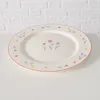 A white oval plate with a floral pattern around the rim and a small cluster of flowers in the center, placed on a white surface against a white brick wall. Produktbild-Vorschau