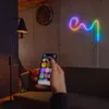 A person is using a smartphone to control a colorful neon light strip in a living room setting. Produktbild-Vorschau