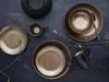 A flat lay of gold and black dinnerware and a black candle holder on a dark textured surface. Náhľad obrázku produktu
