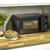 A black microwave oven is shown on a wooden countertop next to a bowl of pasta and a plant. Podgląd zdjęcia produktu