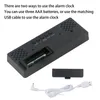 The back of a black digital alarm clock is shown, along with its battery compartment cover and a USB charging cable. Náhled obrázku produktu