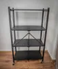 A black metal folding shelf unit with four perforated shelves and wheels, placed against a light-colored wall on a wooden floor. Produktbild-Vorschau