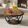 A round coffee table with a wood-look top and a metal base, placed in a living room setting with a fireplace and a chair. Anteprima dell’immagine prodotto