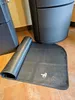 A black mat is partially unrolled on a tiled floor next to a large, dark gray appliance. Náhled obrázku produktu