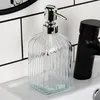 A clear glass soap dispenser with a silver pump sits on a white sink next to a black faucet. Náhled obrázku produktu