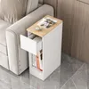 A narrow side table with a drawer and shelves is placed next to a sofa, with magazines and a cup of coffee on top. Náhled obrázku produktu