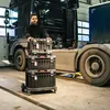 A man stands in a garage or workshop next to a large truck, with a stack of toolboxes on a trolley in front of him. Náhled obrázku produktu