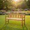 A wooden garden bench sits on a lush green lawn in a garden setting with flowers and a fountain in the background during sunset. Produktbild-Vorschau