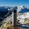 A metal thermos bottle is placed on a rock in a snowy mountain landscape. Produktbild-Vorschau