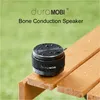 A small, black, cylindrical speaker with water droplets on it, resting on a wet wooden bench. Podgląd zdjęcia produktu