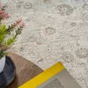 A close-up view of a beige vintage-style rug with a floral pattern, partially visible wooden table, a vase with pink flowers, and a yellow book. Produktbild-Vorschau