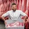 Mann / Metzger mit grauer Plastikkiste mit Fleisch in den Händen, dahinter hängendes Fleisch Produktbild-Vorschau