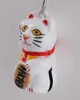 A close-up studio shot of a white lucky cat charm with red and yellow accents, holding a gold oval object with black text. Produktbild-Vorschau