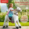 A young girl is happily riding a white mechanical horse toy outdoors, with lush green foliage in the background. Produktbild-Vorschau