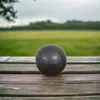 A dark gray ceramic ball sits on a wooden table in an outdoor setting with a blurred green field and trees in the background. Produktbild-Vorschau