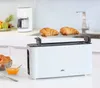 A white BRAUN toaster with two croissants on a warming rack, placed on a kitchen counter with a coffee maker and fruit in the background. Náhled obrázku produktu