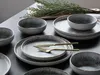 A set of stoneware plates and bowls with a rustic, volcanic-inspired design, arranged on a dark surface with a sprig of greenery in the background. Náhľad obrázku produktu