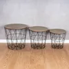Three wire basket side tables with wooden tops are arranged in a row against a concrete wall and wooden floor. Produktbild-Vorschau