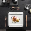 A square white plate with food on it, set on a black wooden table with cutlery and glasses. Produktbild-Vorschau