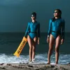 Two women in blue swimsuits stand on a beach with waves at their feet. One woman holds an orange flotation device. Podgląd zdjęcia produktu