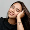 A young woman with long brown hair rests her head on her hand, smiling gently. She is wearing a black t-shirt and a silver bracelet with a red charm. Produktbild-Vorschau