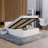A modern white bed with its storage compartment open, revealing the slatted base, in a bedroom setting with wooden wall paneling and a grey rug. Produktbild-Vorschau