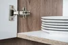A close-up view of a cabinet hinge and a stack of white plates on a shelf. Produktbild-Vorschau