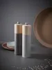 Two salt and pepper grinders on a dark surface with plates and a large platter in the background. Produktbild-Vorschau