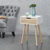 A bedside table with a drawer and wooden legs is placed next to a grey armchair in a room with a textured grey wall and concrete floor. Náhled obrázku produktu