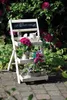 A white, three-tiered plant stand with potted flowers and decorative items is set up outdoors on a brick patio, surrounded by lush green foliage and pink rhododendron flowers. Produktbild-Vorschau