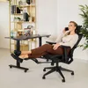A woman is sitting in an ergonomic office chair with her feet up on a footrest, talking on her phone. The chair is next to a height-adjustable desk with a laptop on it. A bookshelf is in the background. Produktbild-Vorschau