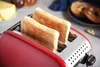 A close-up shot of a red toaster with two slices of toast inside, set against a light gray and white background with a block of butter and some bagels visible in the background. Náhľad obrázku produktu