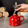 A hand is inserting a metal wick holder into a red and white polka dot ceramic oil lamp. A glass chimney is to the left, and a bowl of oranges is in the background. Náhled obrázku produktu