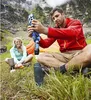 A man and a woman are sitting on grass in a mountainous area, with a tent in the background. The man is demonstrating how to use a water filter. Produktbild-Vorschau