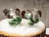 Two clear glass Christmas ornaments filled with pinecones and faux pine branches, resting on a snowy surface atop a wooden log slice, with a textured gray background. Podgląd zdjęcia produktu
