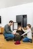 A father and son are by a fireplace, with the son holding a stick and the father putting logs into a basket. Produktbild-Vorschau