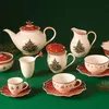 A collection of Christmas-themed tableware, including a teapot, creamer, mugs, and saucers, arranged on a green surface. Anteprima dell’immagine prodotto