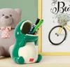 A ceramic dinosaur pen holder sits on a shelf next to a teddy bear and a framed picture of a bicycle. Produktbild-Vorschau