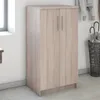 A light brown wooden cabinet with two doors and silver handles, standing on a light gray floor against a white wall. An umbrella stand with umbrellas is visible to the right. Podgląd zdjęcia produktu
