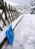 A blue snow shovel leans against a wooden fence next to a snow-covered path. Produktbild-Vorschau