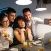 Four young adults are taking a selfie in a dimly lit room, with drinks and a laptop on the table. Voir les images du produit