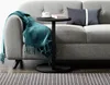 A black C-shaped side table with a teal throw blanket draped over a gray sofa, a white cup and saucer, and a book on top. Voir les images du produit